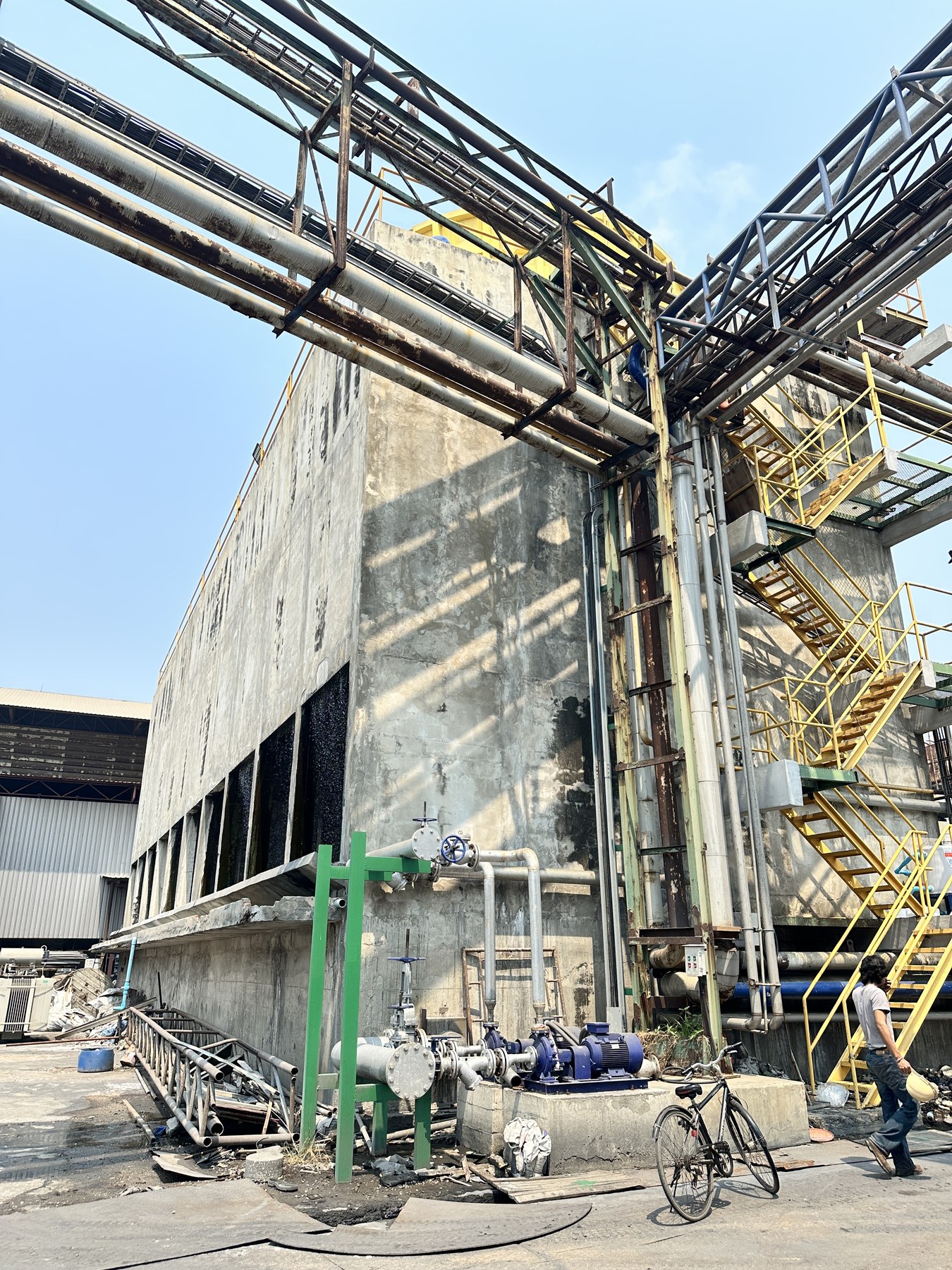 Concrete cooling tower at the Ratchaburi site seen from ground level, with riser pipework, valves and circulating pump alongside the air-inlet openings